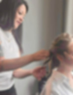 Coiffeuse tressant les cheveux d'une femme blonde dans une pièce grise.