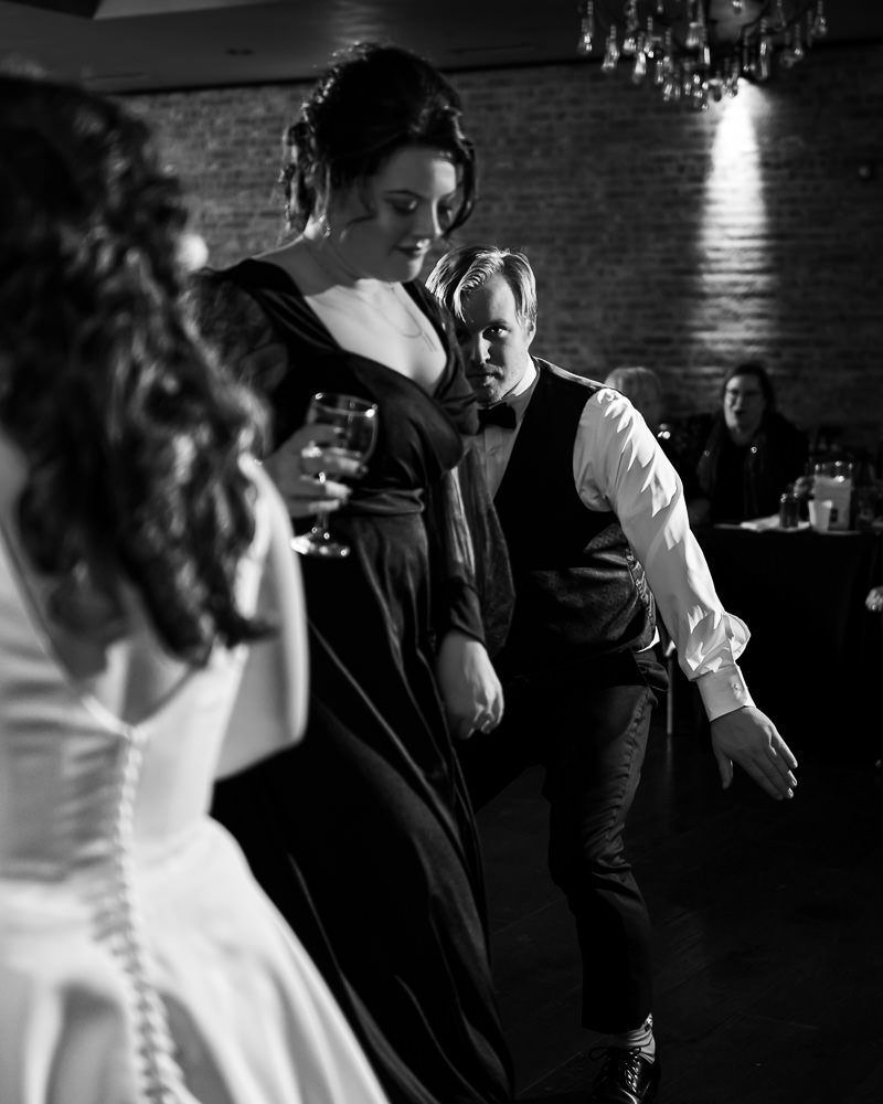 Black and white photo: Wedding guests are captured in motion, enjoying themselves while dancing at the reception.