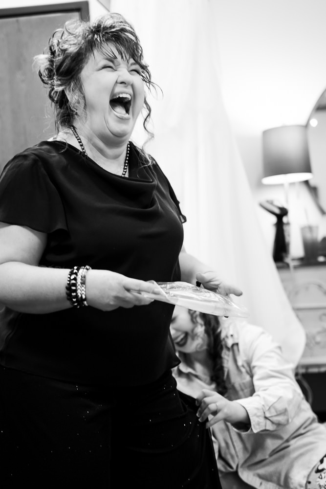 Black and white photo: The bride and her mother share a lighthearted moment, both laughing as they adjust the mother's outfit.