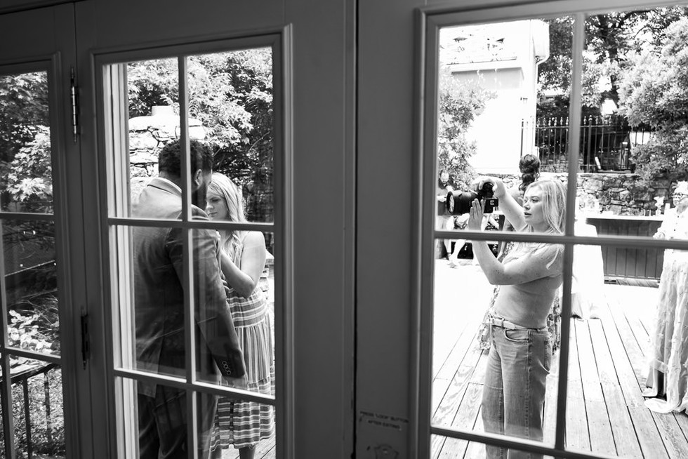View through sunroom window: photographer shooting while planner adjusts groom’s suit