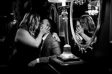 A couple during their engagement session in a restaurant in Kansas City is kissing romantically. A mirror reflects their kiss.