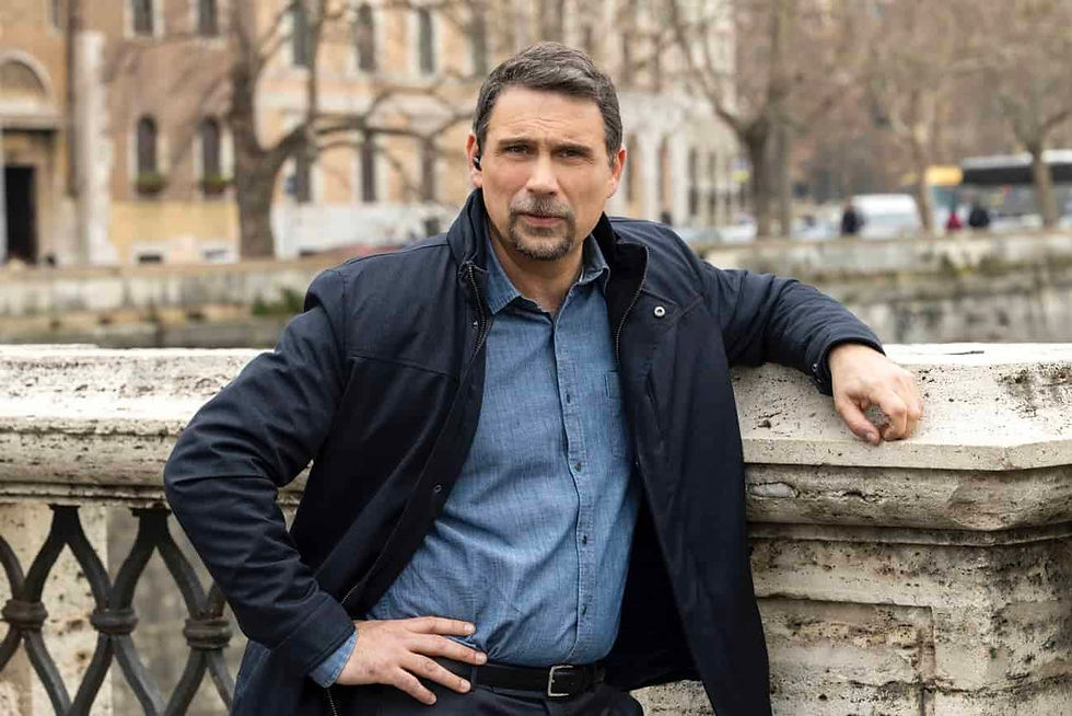 Man in a blue shirt and black jacket leans on a stone bridge railing. Urban background with trees and buildings. Neutral expression.