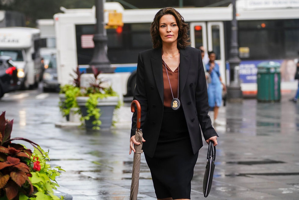 Woman in a black suit walks confidently on wet city street, holding an umbrella and briefcase. Background shows buses and plants.