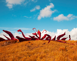 This is a colourful landscape photograph of an orange mountainside of grass set against a bold blue sky with small white clouds in the distance. In the centre of the image stands a woman with 15 lengths of fabric flying up in the air all around her.