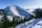 Snow-covered mountain with pine trees, clear blue sky, and misty clouds in the background creating a serene, wintery scene.