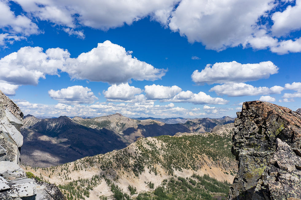 Rocky mountain landscape under a bright blue sky with scattered fluffy clouds. The rugged terrain is dotted with patches of green vegetation.