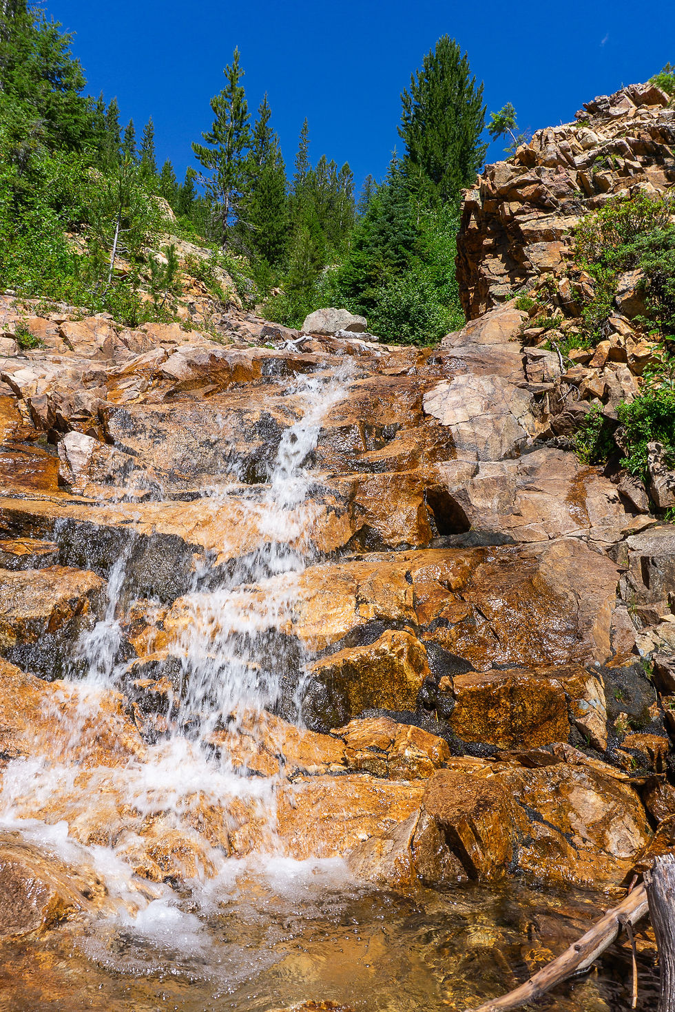 Water cascades over brown rocks, surrounded by lush green trees and under a vibrant blue sky, creating a serene natural scene.