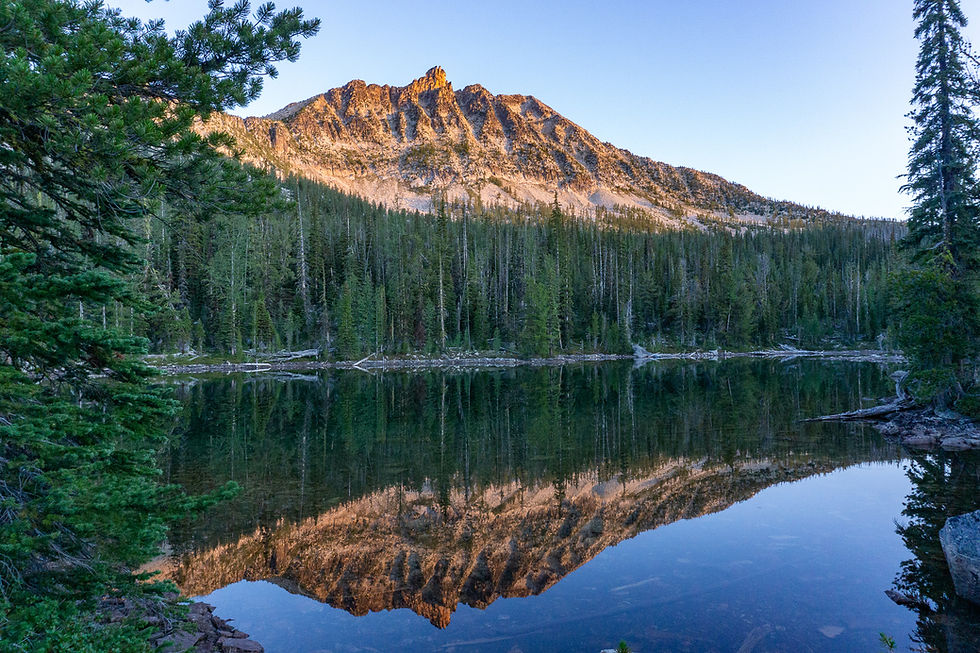 Mountain peak at sunset reflects in a calm lake, surrounded by evergreen trees. Clear sky enhances the serene, natural scene.
