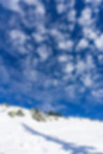 Snowy hill with rocks and conifer trees under a vibrant blue sky filled with scattered clouds. Shadow of trees stretches across the snow.