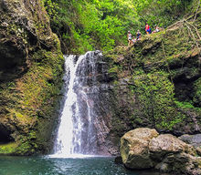 Low-Angle shot of a hiking group standing on top of another lower tier level Makamaka'ole waterfall cascading into a pool