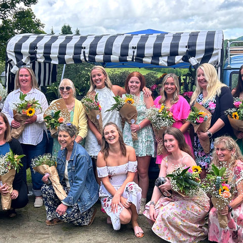 Group of smiling women holding flower bouquets outdoors in front of Ringo the Flower Truck a 1962 VW available for rent.