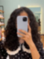 Person with curly hair takes a mirror selfie in a salon, wearing a black polka dot dress. Phone obscures face. Bright, modern background.
