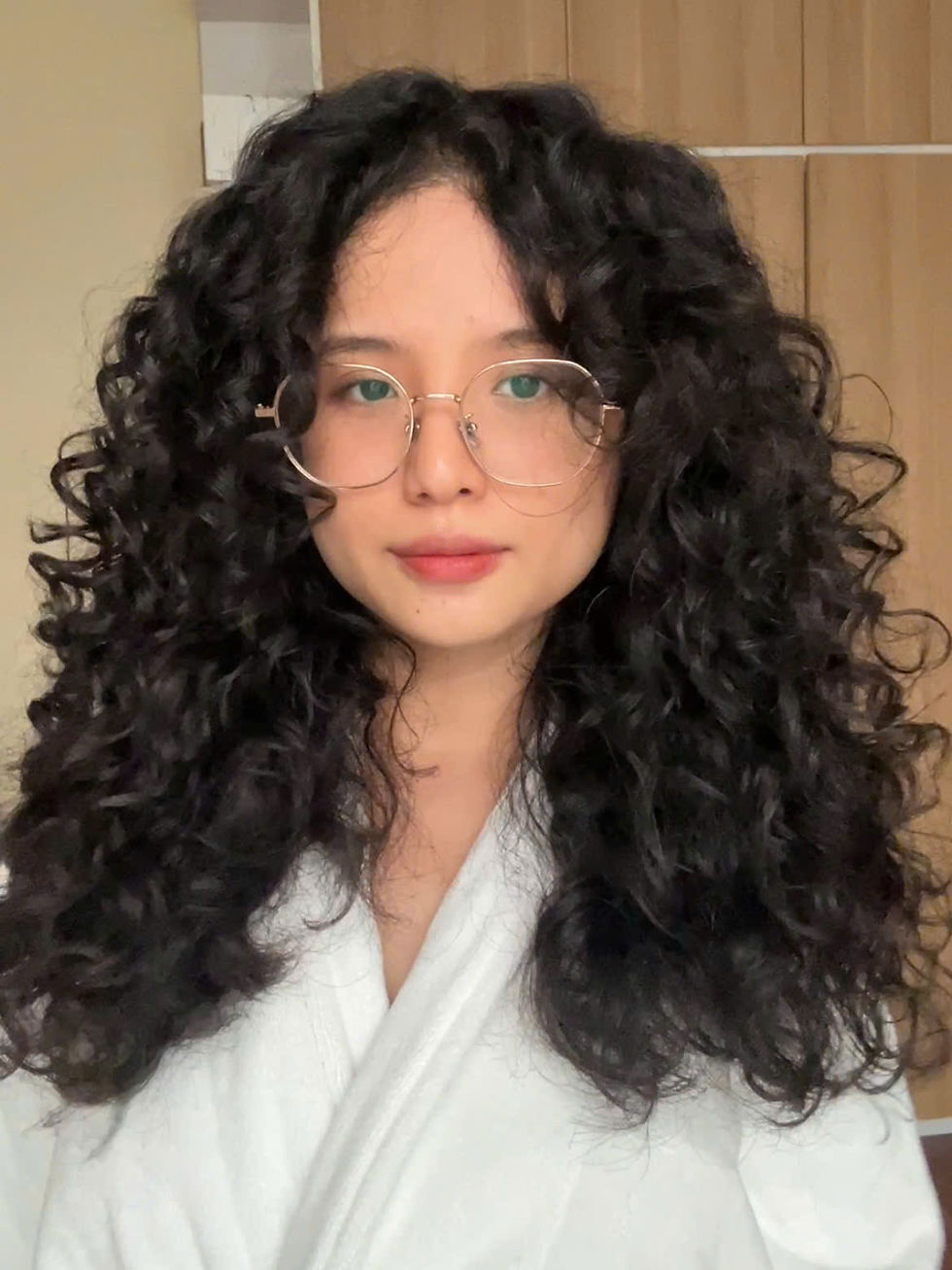 Woman with curly hair and glasses, wearing a white robe, stands indoors with a neutral expression. Wardrobe in the background.