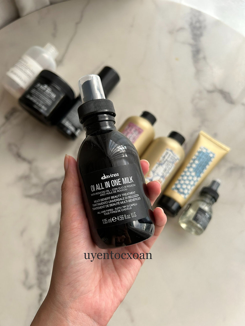 Hand holding a black Davines OI All In One Milk spray bottle over a marble counter with other haircare products; uyentocxoan