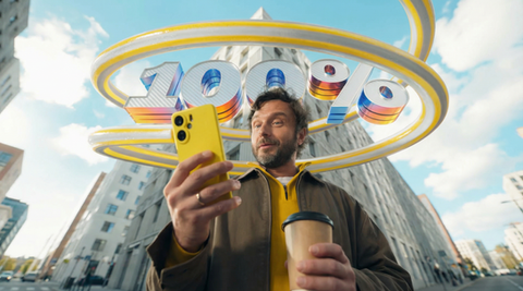 A bearded man in a jacket takes a selfie with a yellow smartphone while holding a takeaway coffee cup; behind him, large shin