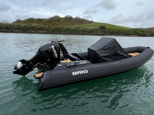 Used ribs for sale - a Black BRIG Eagle 6.7 Custom inflatable boat with Suzuki motor floats on calm water, tied to orange buoys. Green hills and cloudy sky in the background.