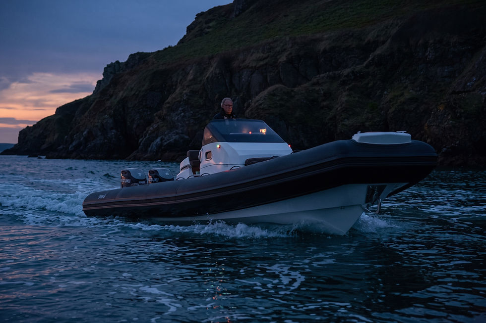 RIB motorboat with a man aboard speeds past dark cliffs at dusk, Suzuki outboards visible.