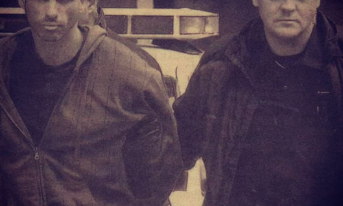 Two men, one handcuffed, walk with a police car in the background. It's a sepia-toned image, conveying a serious mood.