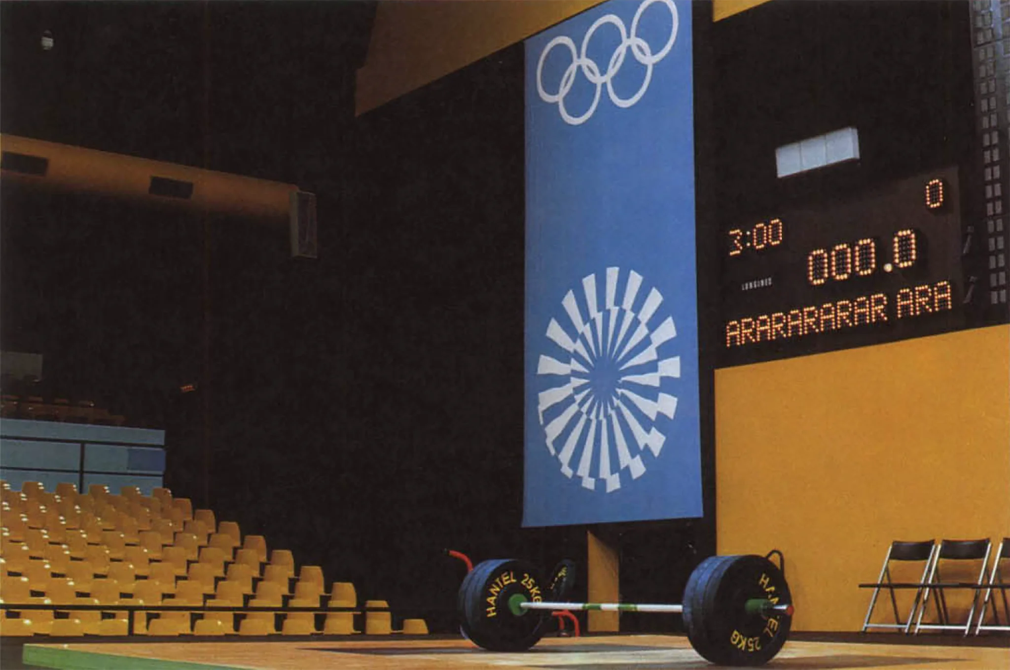 Empty weightlifting stage with a barbell, Olympic rings banner, and digital scoreboard. Yellow seating contrasts with a dim setting.