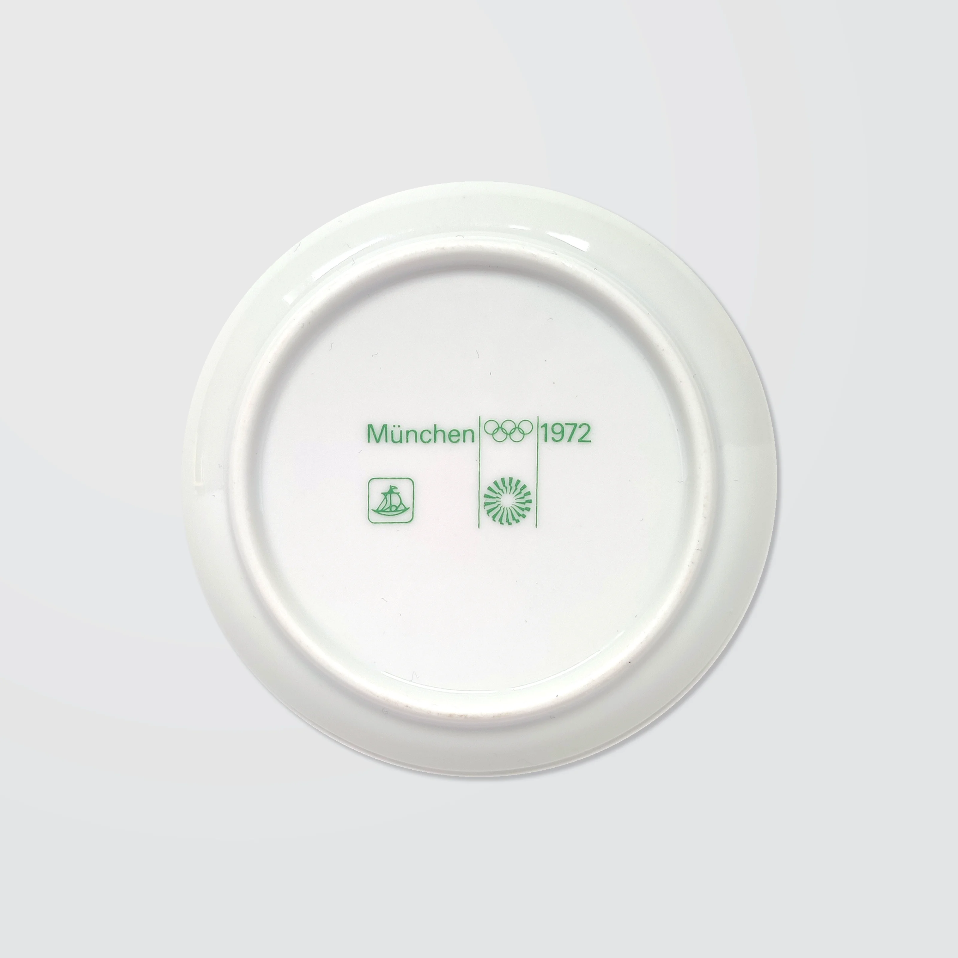 White ceramic coaster with green text: "München 1972," Olympic rings, and logos. Clean, minimalist design on a light grey background.