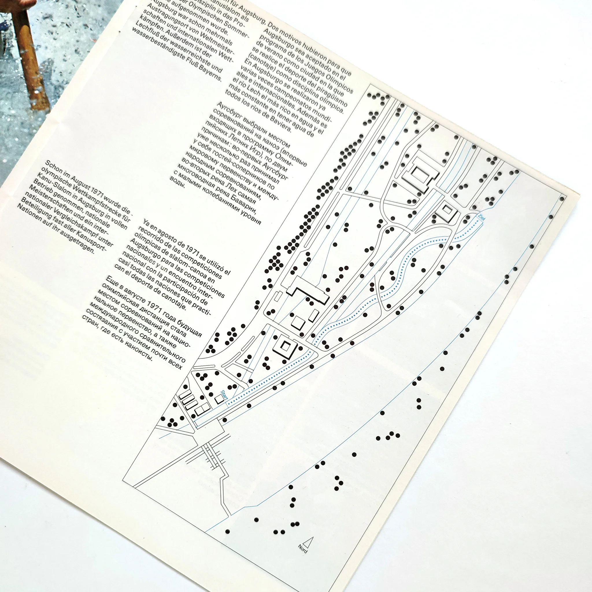 Map with dotted paths and structures. Text in multiple languages. Light background, blue + black lines highlight details.