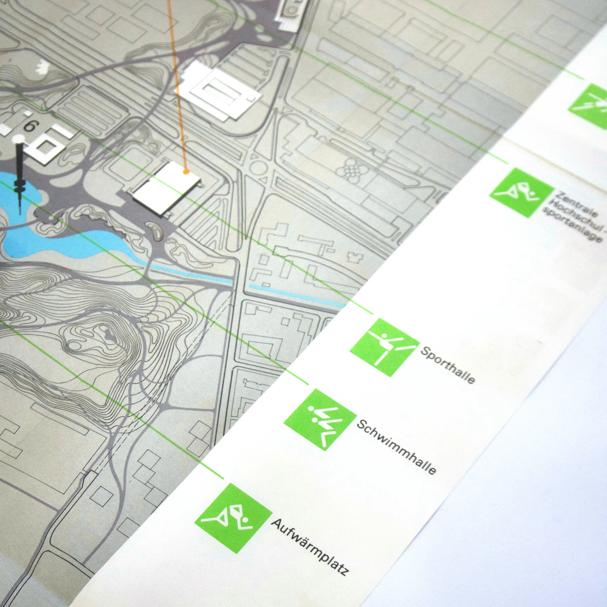 Map with a sports complex layout, featuring paths, water, and contour lines. Green icons represent locations: Sporthalle, Schwimmhalle, Aufwärmplatz.