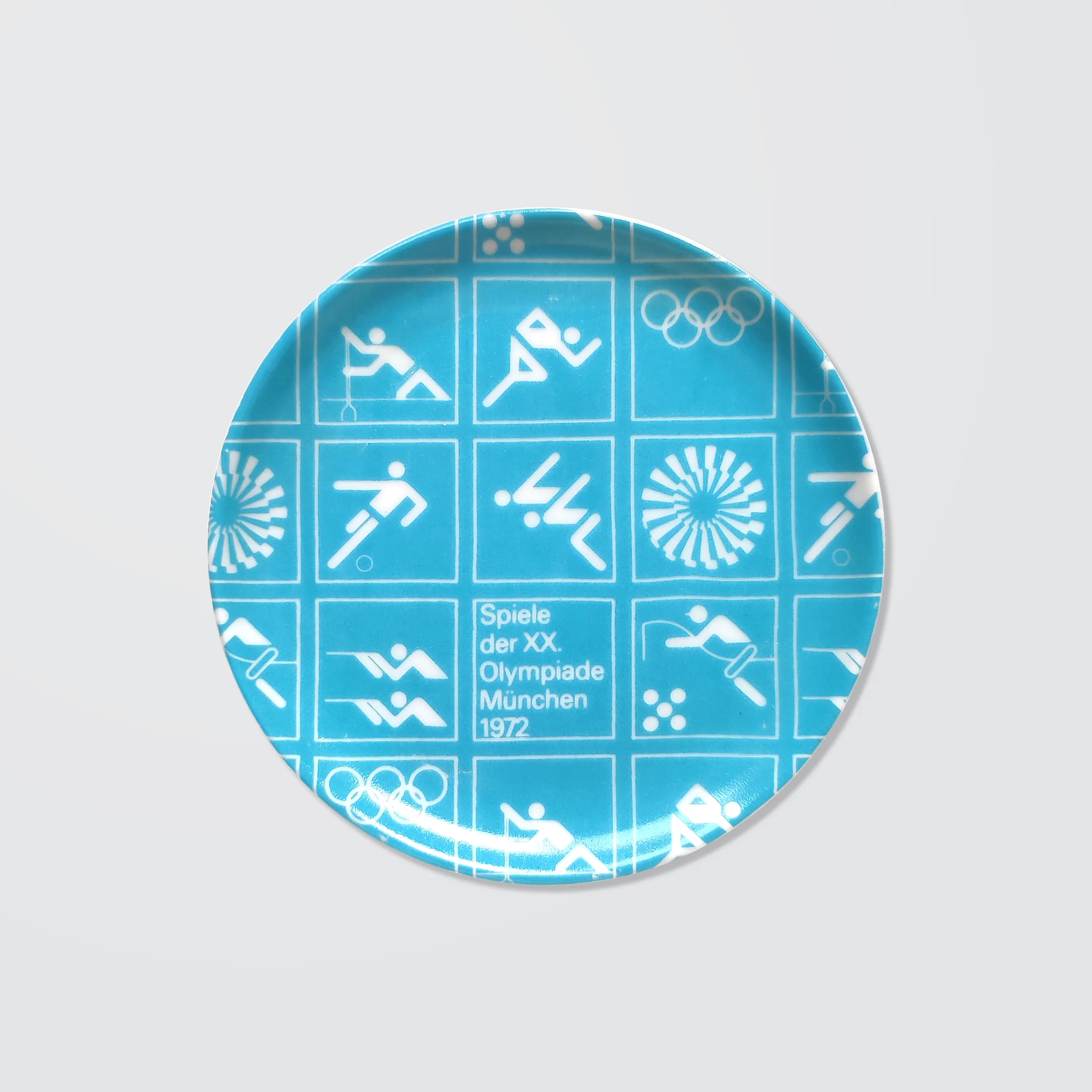 Light blue ceramic coaster with white Olympic symbols from the 1972 Munich Games, including athletic icons and "Spiele der XX. Olympiade München 1972".