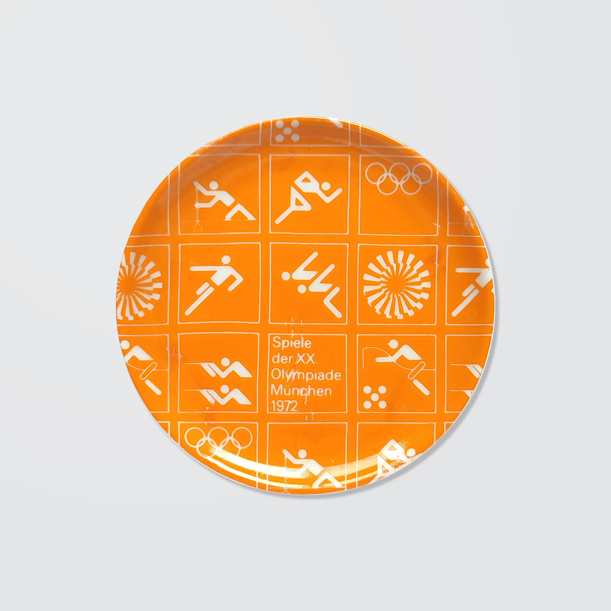 Orange ceramic coaster with white Olympic symbols from the 1972 Munich Games, including athletic icons and "Spiele der XX. Olympiade München 1972".