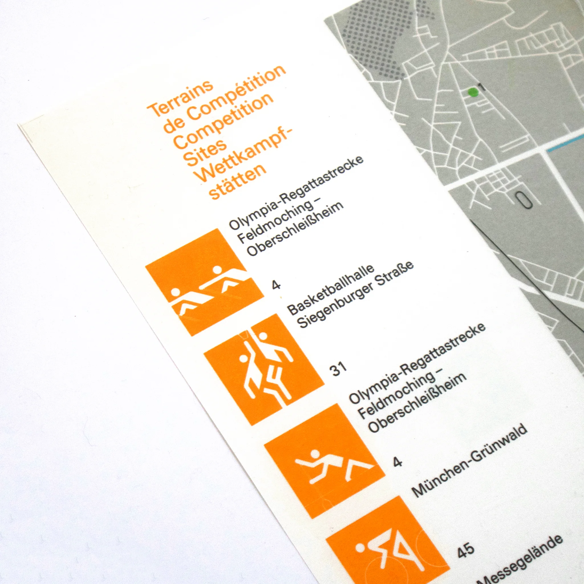 Map close-up showing competition sites with orange sports icons. Text in multiple languages, partially visible map in grey on the right.