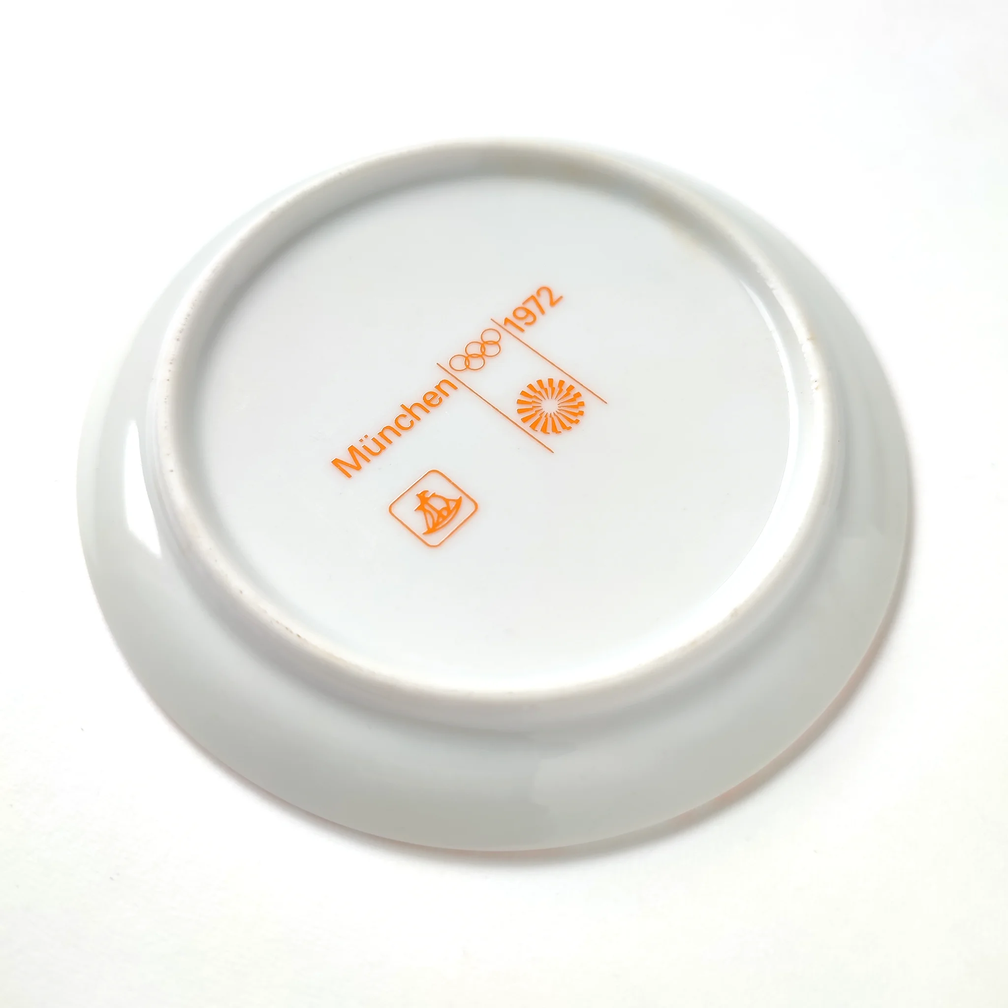 White ceramic coaster with orange text: "München 1972," Olympic rings, and logos. Clean, minimalist design on a light grey background.