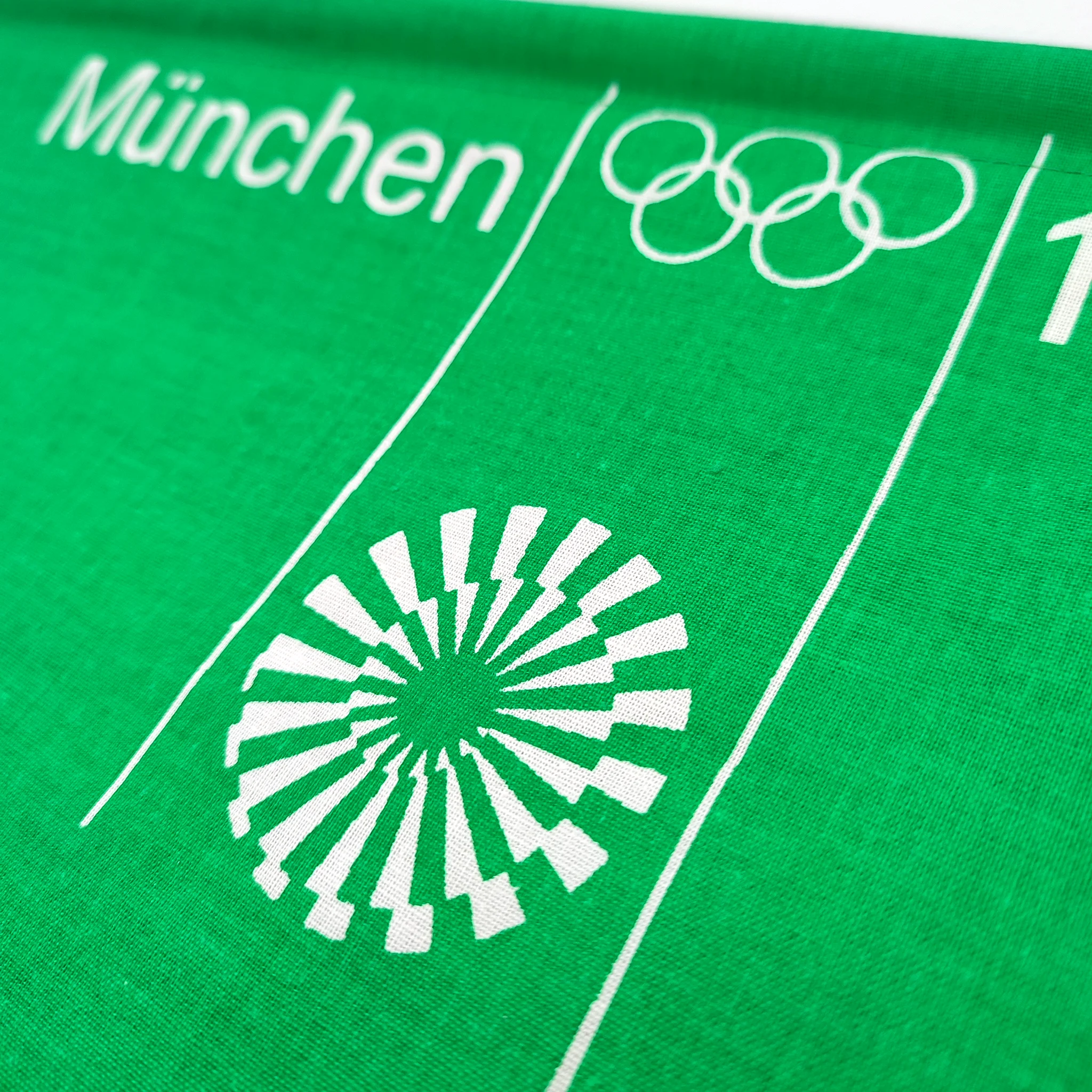 Green fabric featuring white text "München", Olympic rings, and a geometric sunburst design, suggests an Olympic theme.