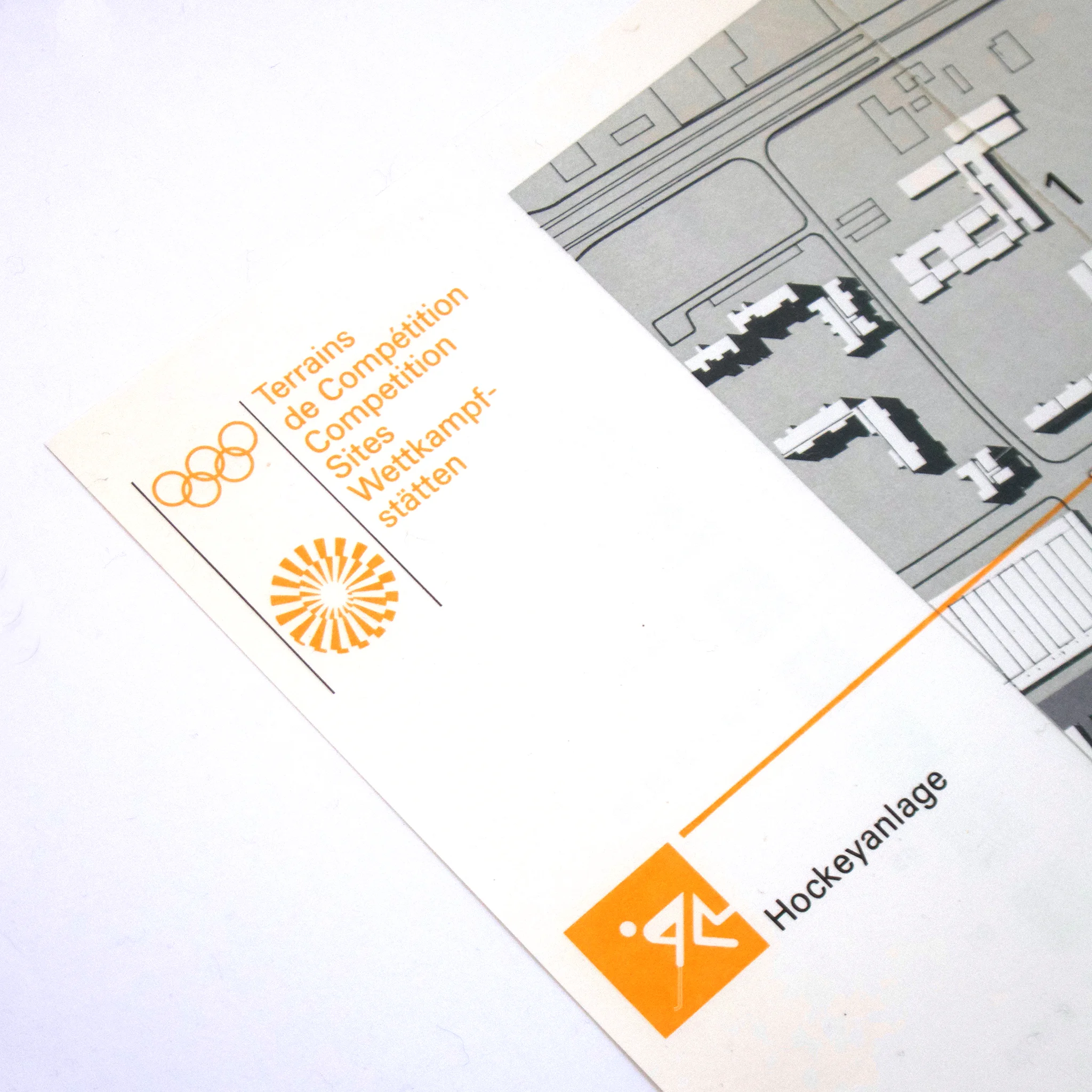 Map detail with multilingual text, Olympic rings, orange pattern, and labeled "Hockeyanlage." Grey and white map layout.