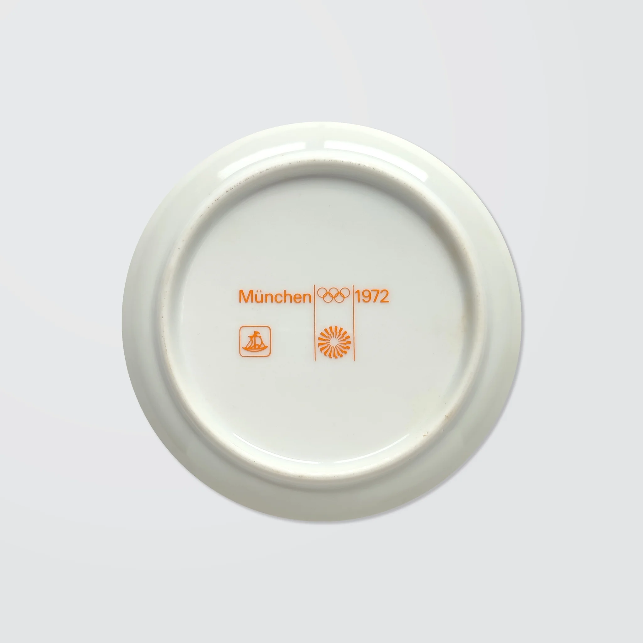 White ceramic coaster with orange text: "München 1972," Olympic rings, and logos. Clean, minimalist design on a light grey background.
