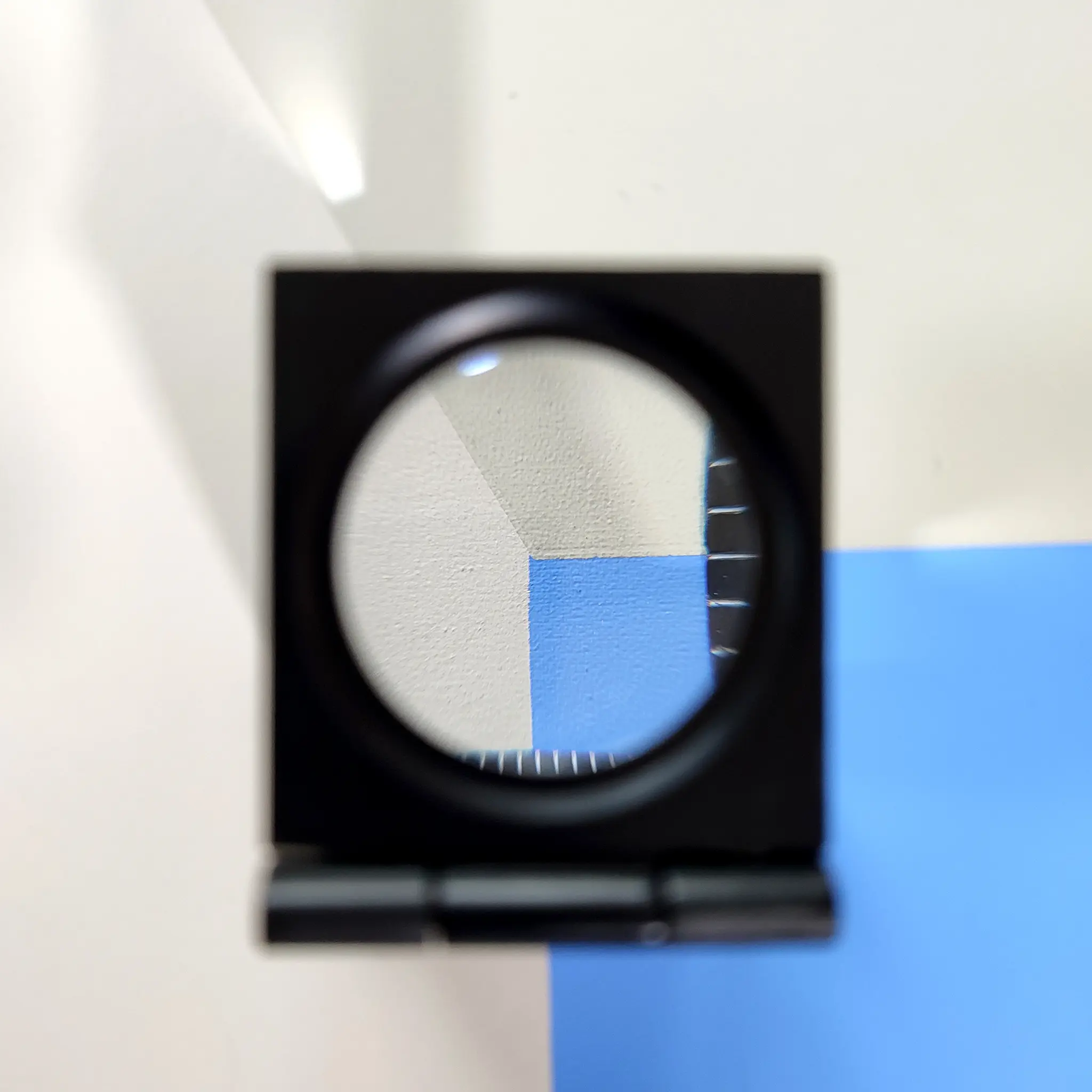 Black square magnifying lens over a beige and blue geometric pattern, highlighting textured surface. Minimalist and abstract setting.