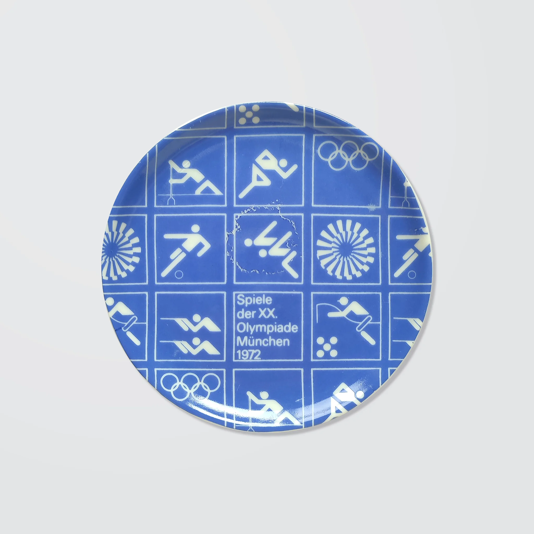 Dark blue/violet ceramic coaster with white Olympic symbols from the 1972 Munich Games, including athletic icons and "Spiele der XX. Olympiade München 1972".