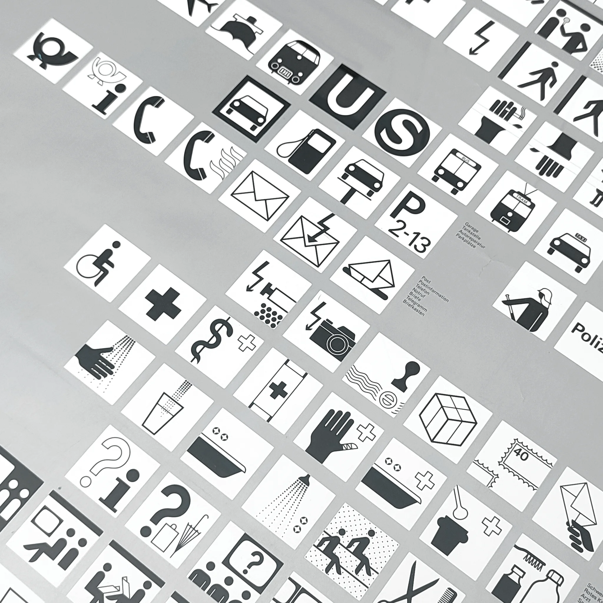 Black-and-white grid of transit and service icons on silver, with letters U and S, bus, tram, taxi, phone, mail, and medical symbols