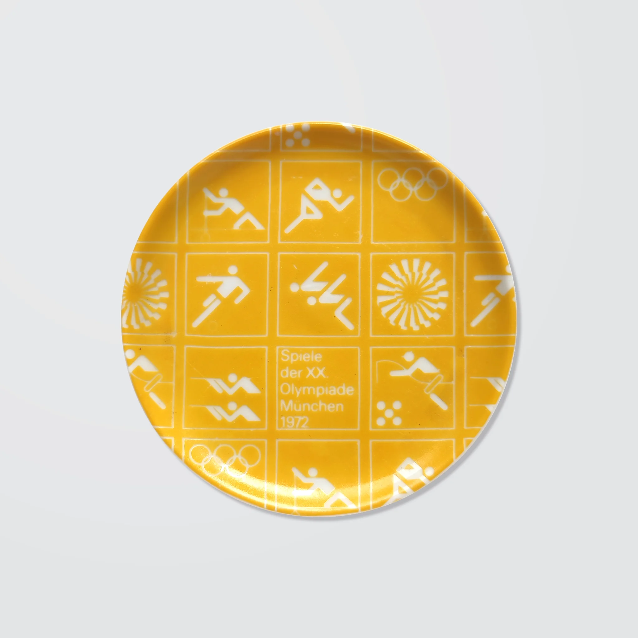 Yellow ceramic coaster with white Olympic symbols from the 1972 Munich Games, including athletic icons and "Spiele der XX. Olympiade München 1972".