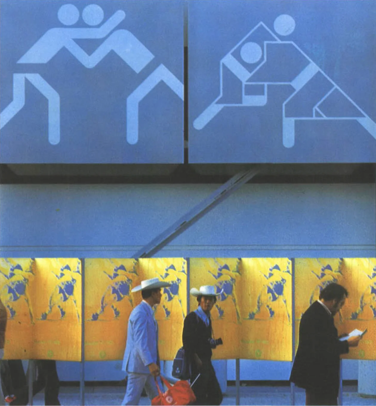 People walk past panels of wrestling posters below two large pictogram signs for the Olympic Wrestling Judo Hall