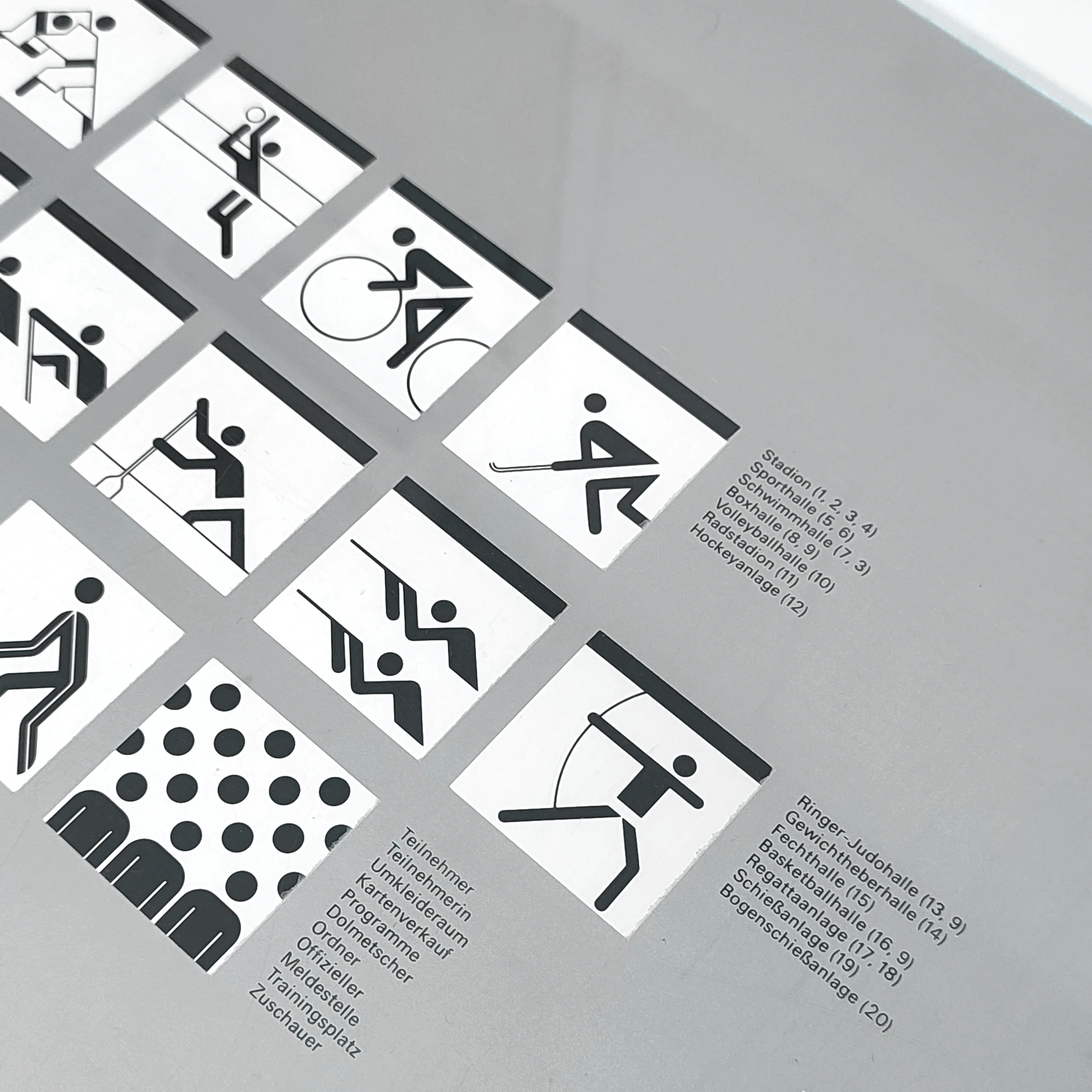 Black-and-white sports pictogram chart with German labels for events like cycling, fencing, archery, and hockey on a silver background.