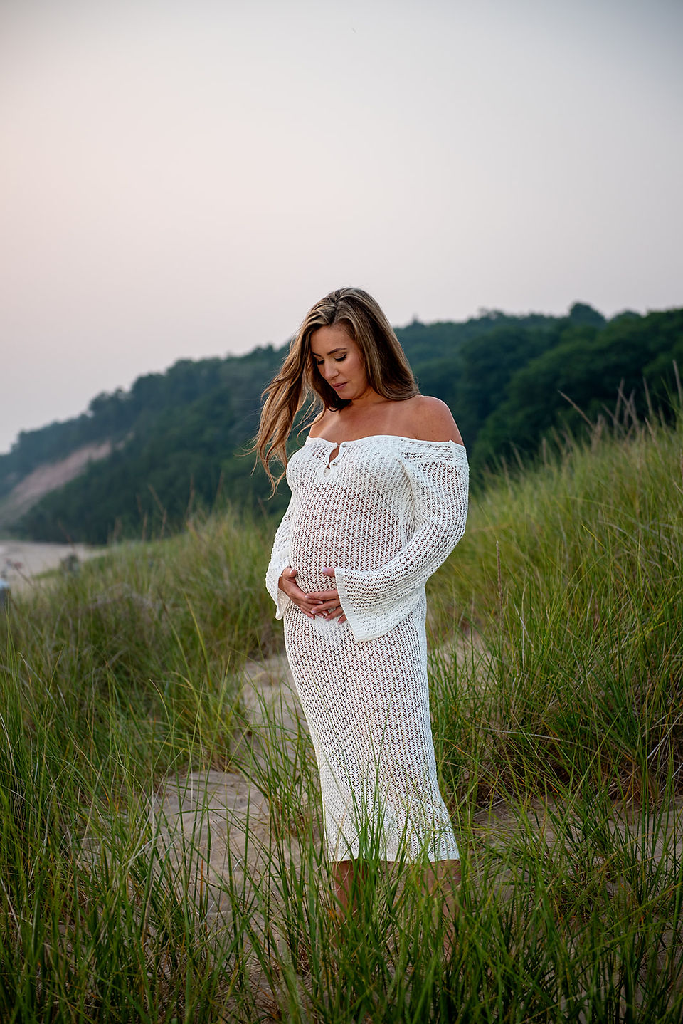 A pregnant woman on a sandy path on the beach in Frankfort Michigan. She is caressing her belly with her right hand while looking down. Her hair is blowing in the wind