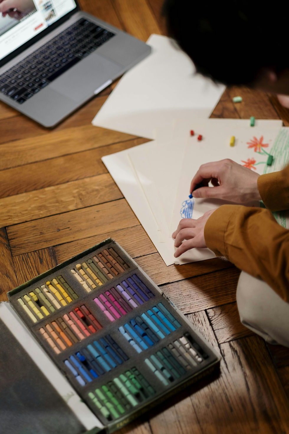 Person drawing with crayons on paper on a wooden floor. A colorful box of pastels and a laptop displaying video are visible. Cozy setting.