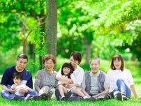 緑の木々に囲まれた公園で、子どもから高齢者まで三世代の家族が芝生に座り、笑顔で寄り添っている様子。