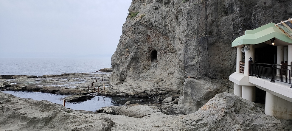 洞窟の入り口付近から見た海辺の景色。手前には波に削られた平らな岩場が広がり、奥には海が広がっている。岩壁の一部に小さな洞穴が見える。