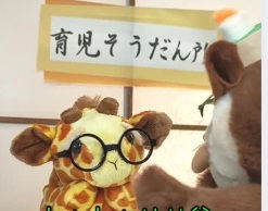 眼鏡をかけたキリンの人形とみみずくの人形が、『育児そうだん所』の看板の前で向かい合って座っている様子。子育て相談をテーマにした場面