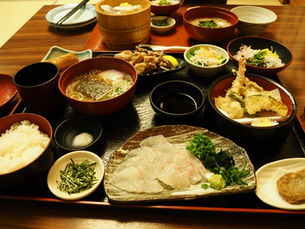 黒いお盆の上に並べられた豪華な和食定食。手前には白身魚の刺身、その隣に天ぷらの盛り合わせ、奥にはそば、ご飯、小鉢などが所狭しと並んでいる。