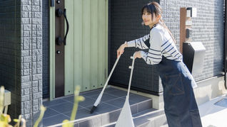 晴れた日の朝、玄関先でホウキとチリトリを持って笑顔で掃除をするエプロン姿の若い女性