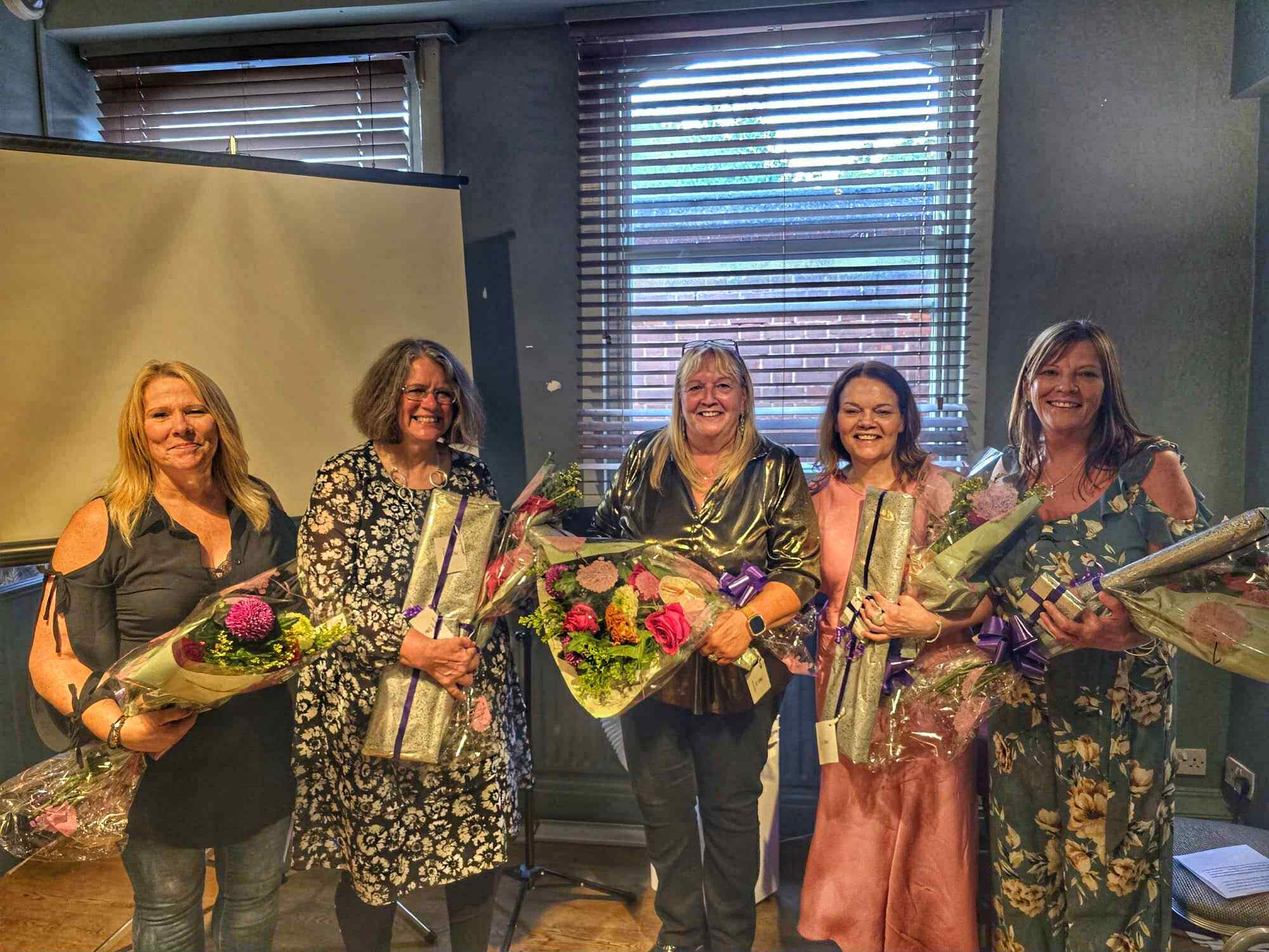 Group of 5 women standing in a row all collecting gifts and flowers.