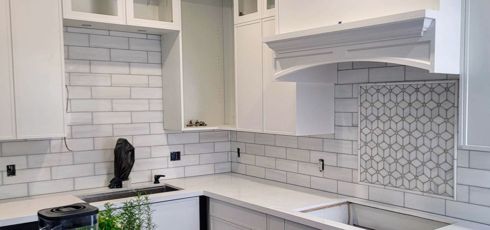 Bright transitional kitchen renovation featuring sleek white cabinetry with minimal hardware, a custom range hood with decorative trim, white subway tile backsplash, a geometric mosaic accent behind the cooktop, and modern quartz countertops.
