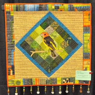 Bird Mosaic is a quilt that features the portrait of a lovebird on a branch.  The bird is surrounded by squares in various shades of green, and this larger square is placed on point.  Twelve beaded tassels hang from the bottom of the quilt.  