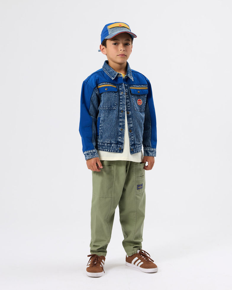 Child standing in full outfit with denim jacket, green pants, and matching cap.